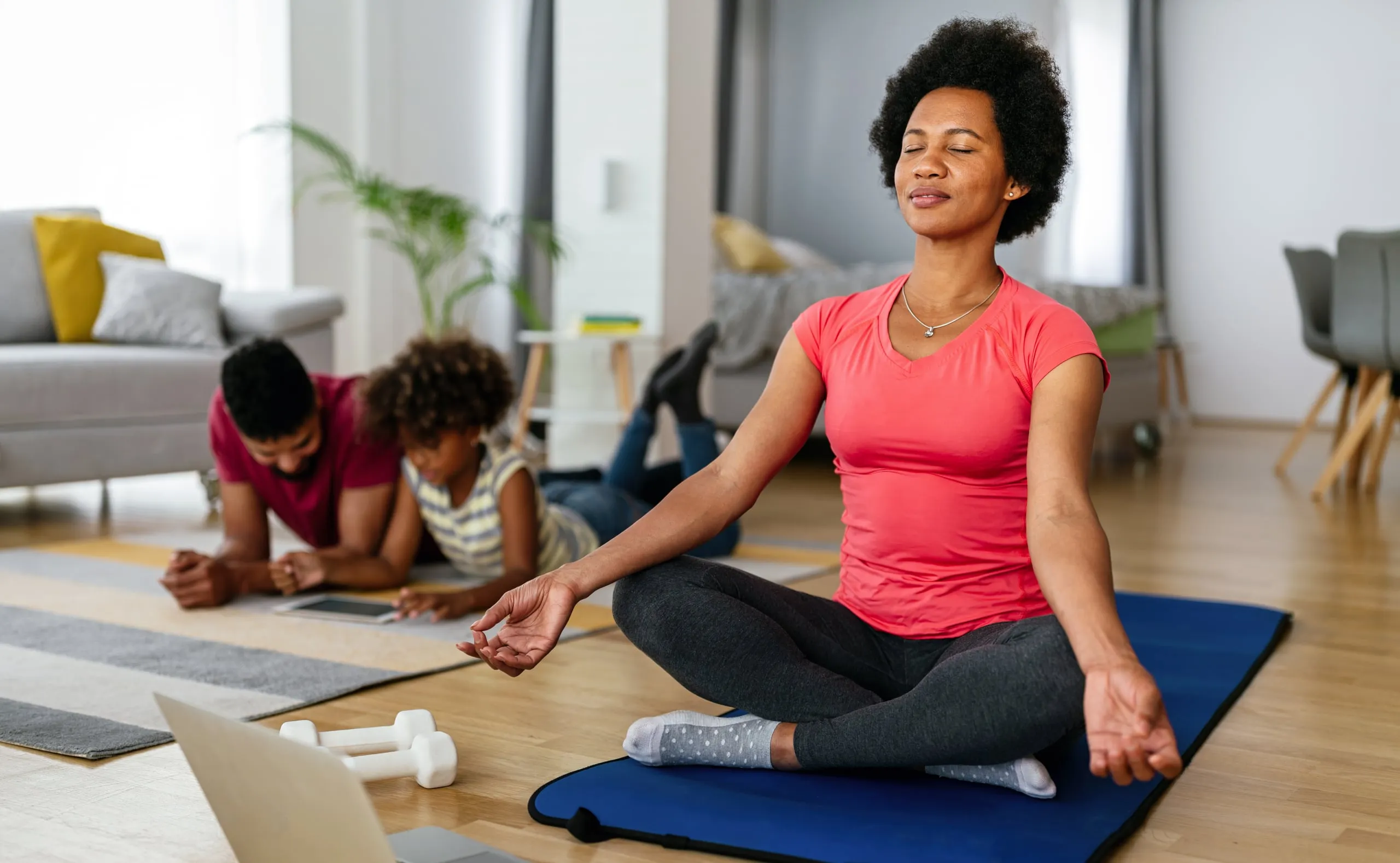 A woman meditating on a mat at home while two children play nearby